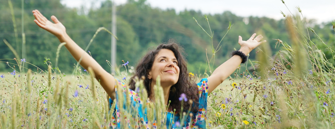 In der Stille erwacht die Freude – Yoga Nidra mit Live-Musik Frau in farbenfroher Kleidung sitzt entspannt mit ausgebreiteten Armen in einer Sommerwiese – Symbolbild für tiefes Loslassen und Lebensfreude beim Yoga Nidra Abend mit Live-Musik bei YOGAKEY.