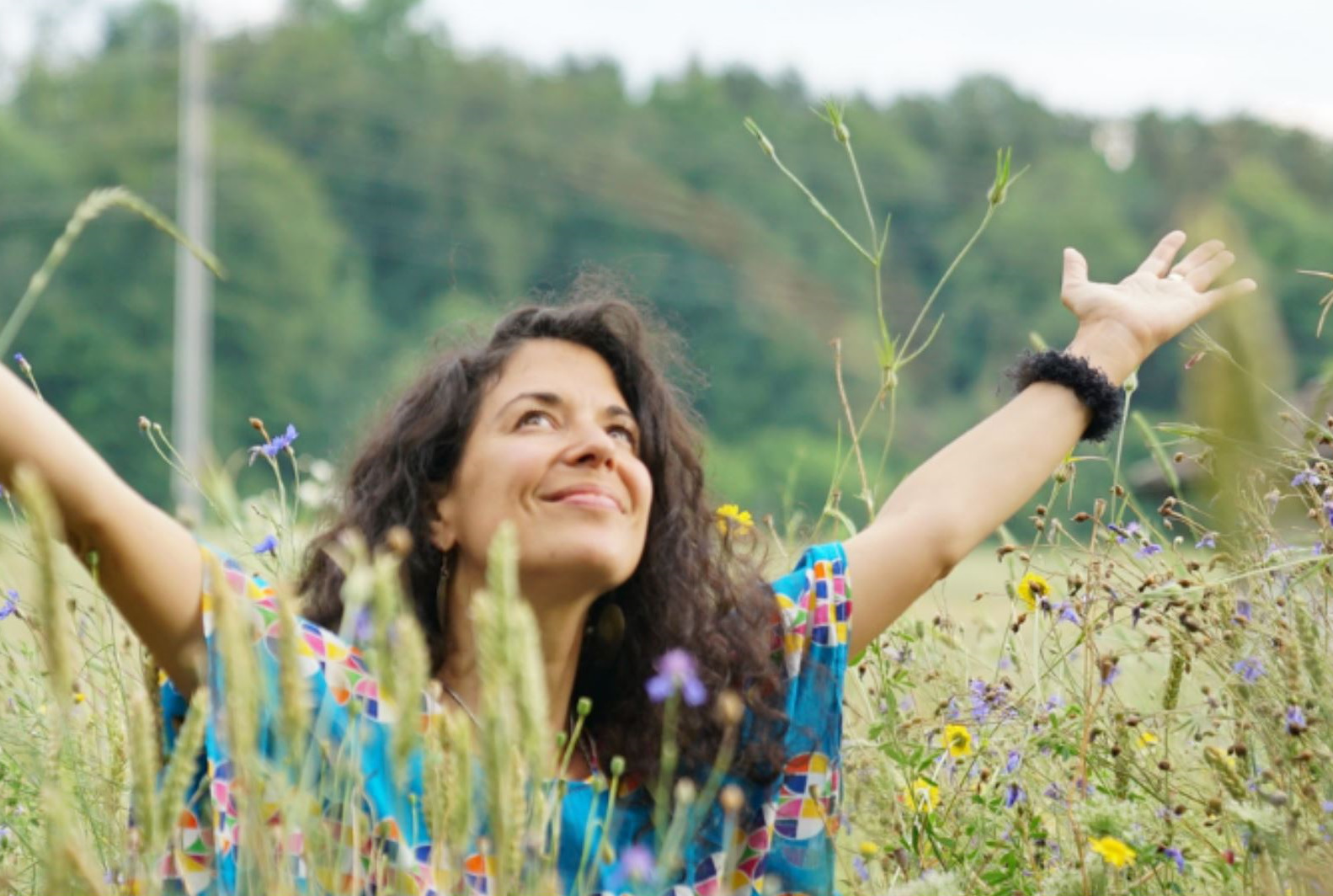 Frau in farbenfroher Kleidung sitzt entspannt mit ausgebreiteten Armen in einer Sommerwiese – Symbolbild für tiefes Loslassen und Lebensfreude beim Yoga Nidra Abend mit Live-Musik bei YOGAKEY.